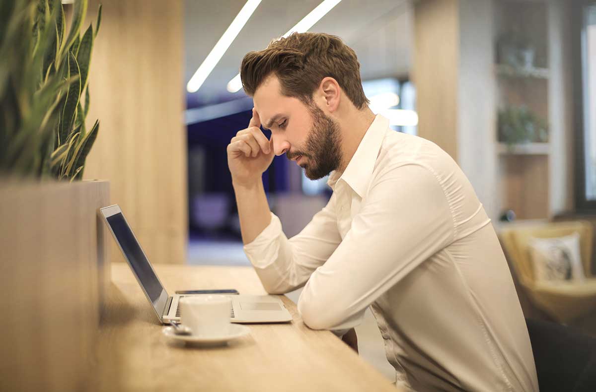 man with headache sitting at desk with laptop and coffee mug