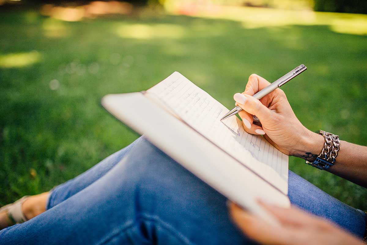 person sitting in grass, writing in journal with silver pen